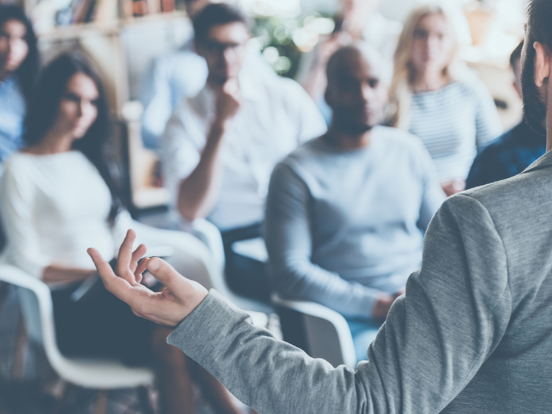 Man standing with back to the camera facing a seated out of focus conference crowd. He is wearing a grey top and has his hands spread as if he is explaining something.