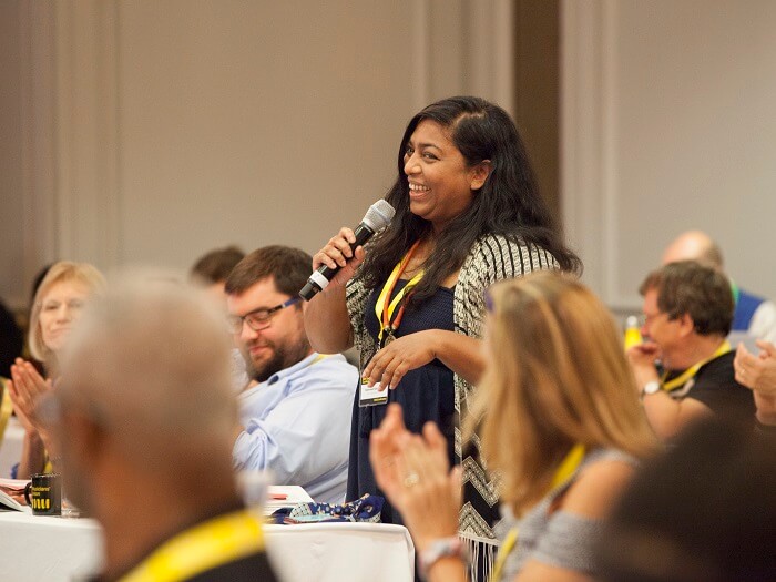 Members of conference smile and clap, while one delegate stands and speaks from the floor.