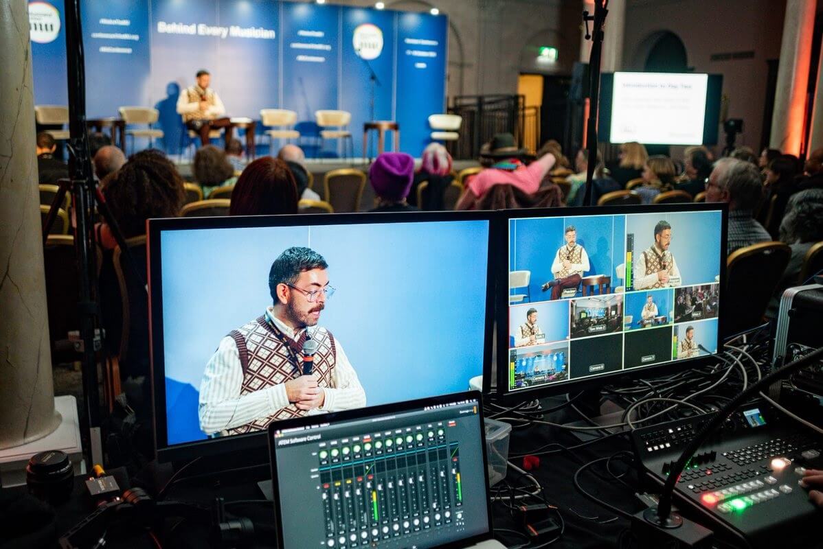 John Shortell speaking at the MU members conference, seen from the perspective of multiple cameras and a sound mixing desk.