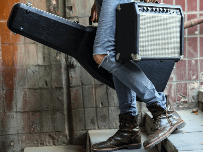 Close up of person in jeans and boots, carrying their guitar case and amp over stone steps.