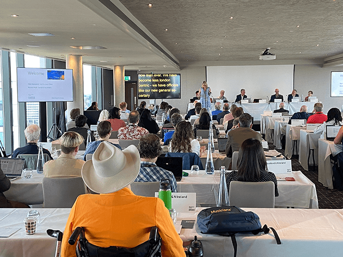 Across a long room full of delegates sat at tables, we see Naomi presenting to the group, speaking into a microphone