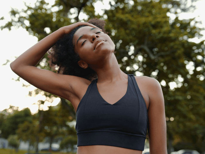A woman sat in a park closing her eyes and gently stretching her neck to the side.