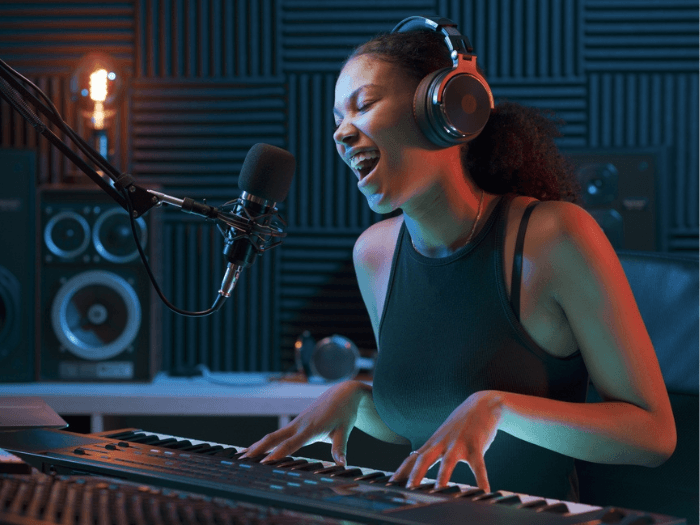 A young female musician in a recording studio playing keyboard and singing into a microphone.