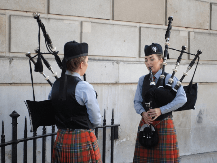 Two young women playing Highland Bagpipes in front of stone wall.